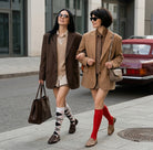 Two women walking on a city street, both wearing brown and beige outfits with patterned socks.